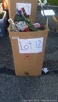 Full view of cardboard box labeled Lot 12 containing Christmas decorations including wrapping paper, greenery, and some ornaments