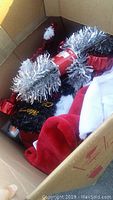 Top view of open box showing silver tinsel garland, red and white Santa hat, black decorative fabric with gold writing, and red box decoration.