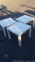 Three white square side tables positioned outdoors on pavement, showing the tops and legs clearly from a slight angle.