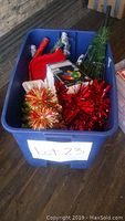 Blue plastic bin filled with assorted Christmas decorations including tinsel pom-poms, mini trees, stocking, Christmas lights and more.