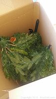 Top view inside the open box showing green artificial tree branches with pinecone decoration and black connector poles for assembly.