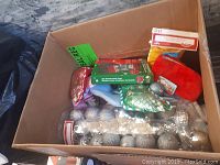 Box filled with various Christmas decorations including silver round ornaments, red stocking, decorative fish ornament, and an ugly sweater bottle cooler.