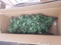 Photo showing one part of the artificial Christmas tree packed inside a cardboard box lying horizontally. The green branches resemble pine needles with some brown stems visible.