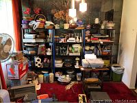 Wide view showing three shelving units fully packed with assorted household items in a cluttered room with carpet and fan