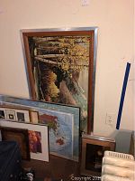 Photo showing a large framed landscape artwork leaning against a wall along with multiple other framed prints including a world map and various sizes of frames stacked together.
