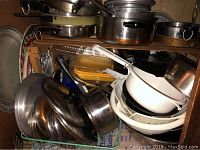 Photo of stacked pots, pans, white and metal colanders, lids, and some kitchen utensils.