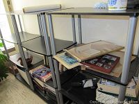 Three black MDF shelving units with metallic supports, showing scratches and used condition. Units have multiple shelves with various items on some shelves.