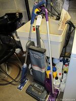 Full view of Hoover Elite II vacuum cleaner, purple floor mop, and cleaning supplies behind them near laundry sink.