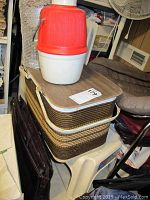 Multi-tiered picnic basket stacked, with red/white beverage cooler on top and white plastic step stool below; partial view of leather or faux leather bag nearby.