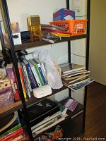 Metal shelving unit with cook books, travel books, magazines, and other small items on multiple shelves. Light bulbs, pouches, and a small basket also visible.