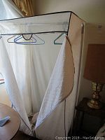 Upright wardrobe rack with fabric zippered cover partially opened, showing hanging rod with assorted hangers and some contents inside.