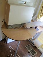 Sewing machine in beige carrying case on top of round wooden sewing table with tripod legs and padded armrest.