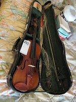 Vintage violin inside black case with green felt interior. Violin shows signs of age and use. Case has compartments and soft lining.