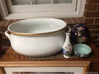 Photo showing large white bowl, blue and gold urn, small white floral vase, and small patterned bowl arranged on wood surface.