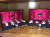 Two black rectangular cases open, each containing 4 silver chalices nestled in red velvet lining.