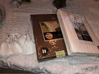 Two packaged tablecloths, one gold Windsor and one white Wasmsutta, displayed on a patterned beige tablecloth with four white tassels to the left.