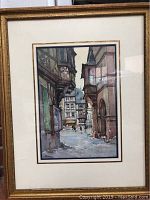 Framed watercolor painting showing a street scene with buildings and figures, signed by the artist.