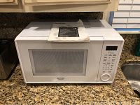 Front and partial top view of Sharp white microwave oven placed on a kitchen counter with the instruction manual on top.