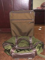 Briggs & Riley chocolate brown carry-on suitcase standing upright behind Banana Republic forest green duffle bag lying down showing leather handles and trim