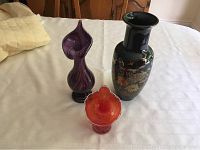 Three vases on white cloth: tall black Asian style vase with colorful floral and bird designs, purple swirled vase with curving pointed top, small orange-red ruffled vase.