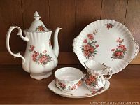 Full view of Royal Albert Centennial Rose 5 piece tea set including teapot with lid, cup, saucer, creamer, and dessert serving plate set against a wood background.