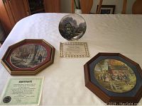 Collector plates on a white tablecloth, two framed plates and one loose plate with certificates of authenticity visible
