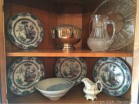 Wood cabinet shelf displaying 4 floral plates, silver plated bowl, glass pitcher, glass tray, decorative bowl, and ceramic creamer.