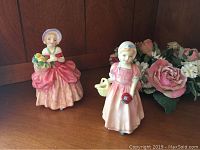 Two Royal Doulton miniature figurines, Cissie and Tinkle Bell, displayed on wood surface next to artificial floral arrangement