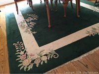 Full view of large wool carpet on hardwood floor showing dark green center with floral border.