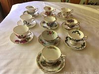 Set of 10 assorted china tea cups with matching saucers showing floral and decorative patterns, laid out on a white cloth surface.