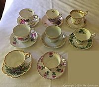 Top view of the eight assorted china tea cups and matching saucers showing patterns and gold trim on white tablecloth.