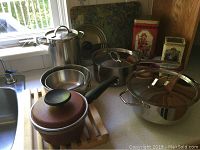 Wide view of assorted cookware on countertop including large stainless steel pots with glass lids, mixing bowls, small saucepan with lid, all arranged near a window.