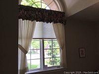 Window treatment with floral valance and sheer white curtains with tiebacks arranged on a residential window showing outside view.