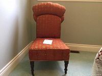 Front view of antique bedroom chair upholstered in patterned reddish-orange fabric, showing distinctive padded backrest and seat, sitting on a light-colored carpet.