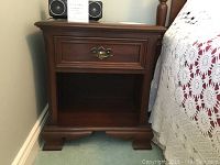 Front view of the Gibbard cherry wood night table positioned next to a bed, showing drawer and open shelf.