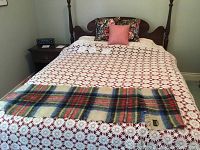 Full bed showing white crocheted bedspread, decorative pillows in floral and pink, and a plaid mohair and wool shawl laid across the foot of the bed.