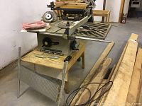 Full view of the Rockwell Beaver table saw on a wooden stand in a basement workshop area with blades and accessories on and around the table.