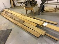 Long, wide, raw wood planks stacked on floor in workshop space with woodworking equipment visible.