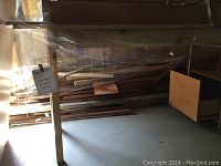 Photo showing long narrow wooden work table with drawers and shelves, placed next to a stack of various sized lumber planks covered with plastic sheeting in a basement.