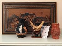 Framed wooden relief art with tropical motif alongside black ceramic vase with human figures, wooden carved stand, and red textured ceramic vessel on a shelf.