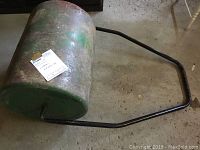 Angled side view of used green metal lawn roller with tubular black handle showing surface rust and chipped paint