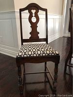 Front view of one dining chair showing dark wood frame with carved detailed back and white cushioned seat with black geometric pattern.