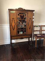 Full view of vintage dining room cabinet with decorative glass door, carved wooden side panels, and turned legs.