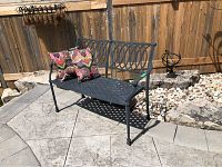 Full view of the black metal garden bench with two patterned pillows placed on it outdoors near rocks and wooden fence.