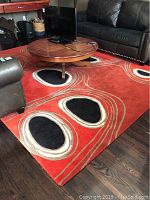 Full view of rectangular red-orange wool rug with abstract black-centered shapes and beige/taupe outlines in living room setting with coffee table and sofas.