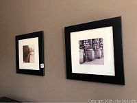 Two black-framed art photographs mounted side by side on a beige wall, showing one photo with umbrellas and the other with a riverside scene.