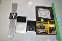 Photo showing two Apple iPods (silver and black) and RhinoShield protective case in original packaging along with a wristwatch with a colorful face.