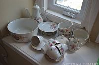 Full set of china items on a windowsill including coffee pot, large bowl, tea cups, saucers, plate, sugar bowl, and creamer with floral patterns.