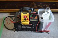 Photo showing Motomaster battery charger, yellow Perry multimeter, black multimeter, and container with battery accessories.