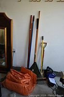 Full group of garden tools standing against wall including rake, broom, and shovels with a blue plastic Kobalt leaf blower and an orange tarp on the ground.